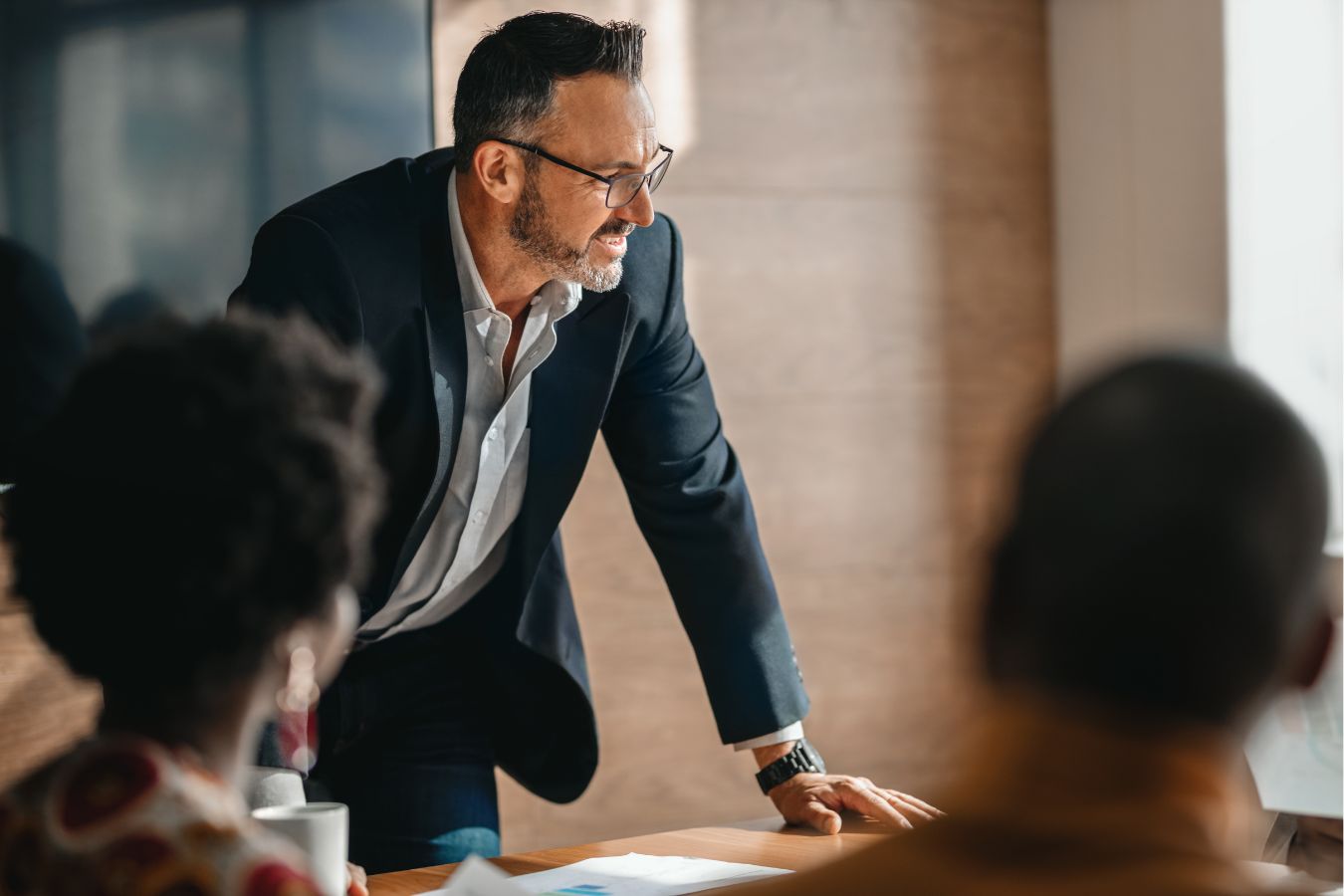 Man in navy suit and glasses leaning over table while talking to others